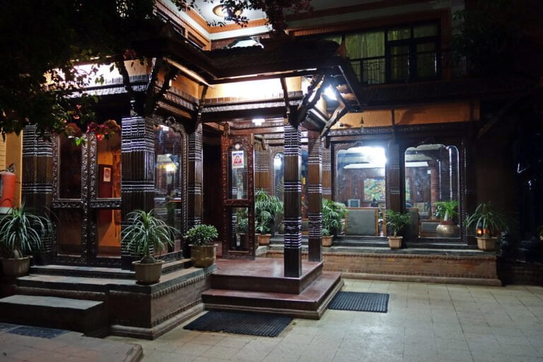An ornate wooden entrance with carved pillars and traditional decor illuminated at night, flanked by potted plants.