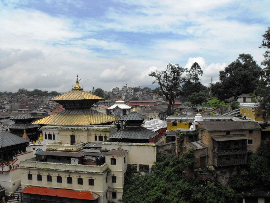 Alt text: A panoramic view of a traditional Nepalese temple complex with distinctive multi-tiered pagoda-style roofs and golden spires, nestled within an urban landscape under a cloudy sky.