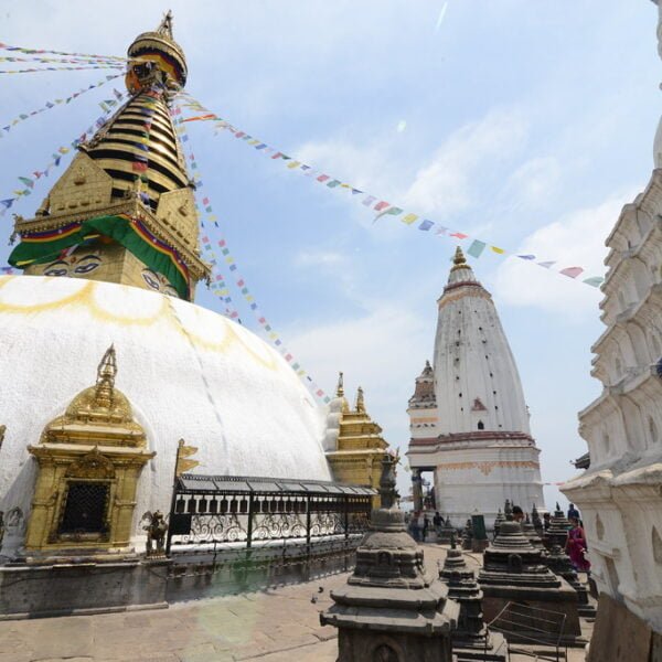 Stupa at Swayambhunath (Monkey Temple) in Kathmandu, Nepal, with a white hemispherical dome and a central spire painted with the eyes of the Buddha. Colorful prayer flags radiate from the top, and smaller shrines and stupas surround it under a clear sky.