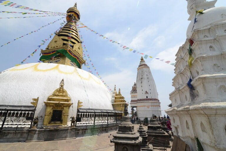 Stupa at Swayambhunath (Monkey Temple) in Kathmandu, Nepal, with a white hemispherical dome and a central spire painted with the eyes of the Buddha. Colorful prayer flags radiate from the top, and smaller shrines and stupas surround it under a clear sky.