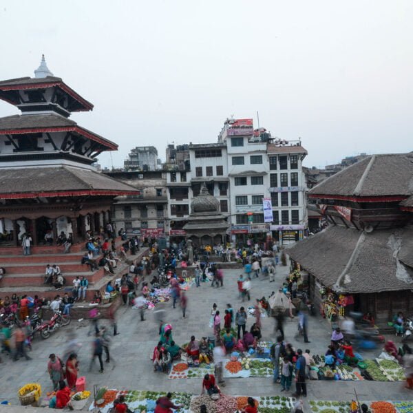 A bustling outdoor market with people gathered around various stalls set against the backdrop of traditional multi-tiered pagoda-style buildings.