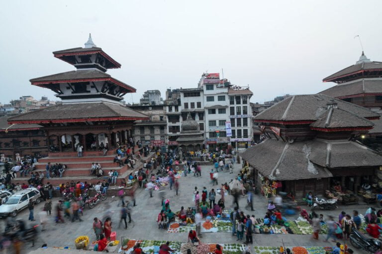 A bustling outdoor market with people gathered around various stalls set against the backdrop of traditional multi-tiered pagoda-style buildings.