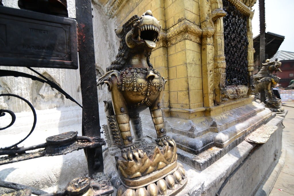 Bronze mythological lion-like statues flanking the corner of a golden religious shrine with intricate designs, set against a backdrop of other architectural elements.
