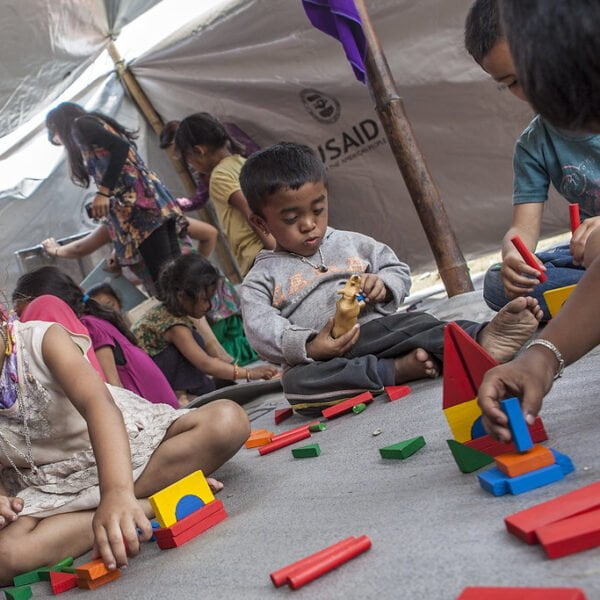 Children playing with colorful blocks on the floor inside a tent with a USAID logo visible.