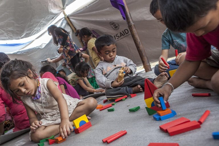Children playing with colorful blocks on the floor inside a tent with a USAID logo visible.