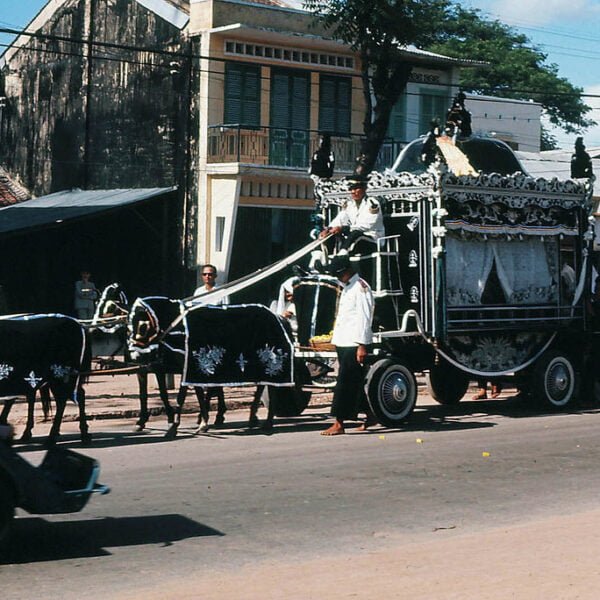 A traditional funeral procession with a decorative horse-drawn hearse on a city street, accompanied by a group of people dressed in white mourning attire.