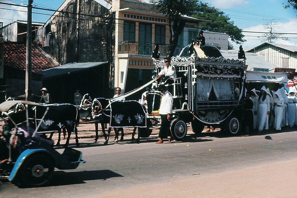 A traditional funeral procession with a decorative horse-drawn hearse on a city street, accompanied by a group of people dressed in white mourning attire.