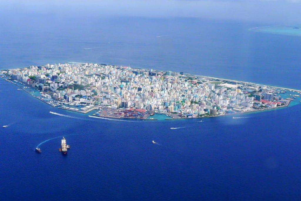 Aerial view of a densely populated island city surrounded by turquoise waters, with boats nearby and a clear view of coastal features.