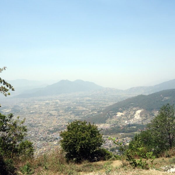 A panoramic view overlooking a valley with scattered buildings, surrounded by mountains under a hazy blue sky, with vegetation in the foreground.