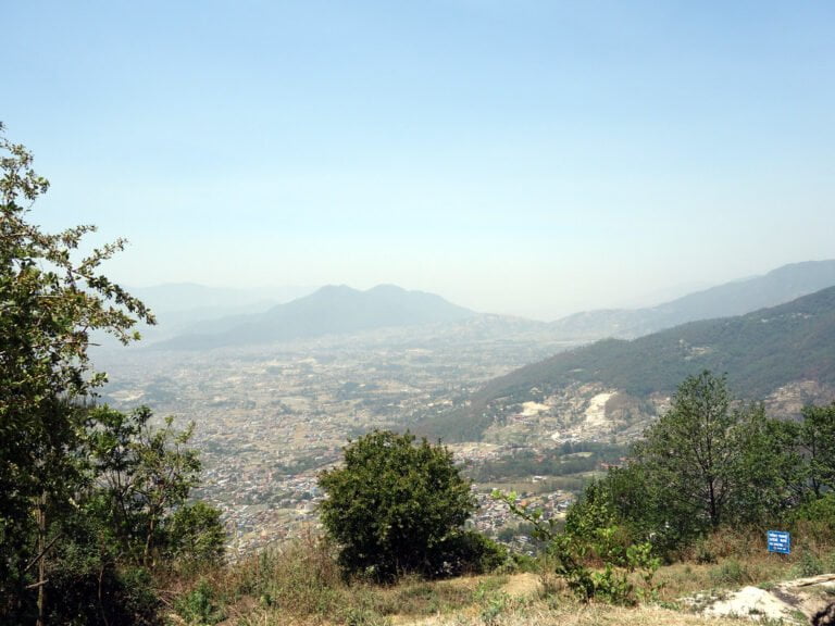 A panoramic view overlooking a valley with scattered buildings, surrounded by mountains under a hazy blue sky, with vegetation in the foreground.