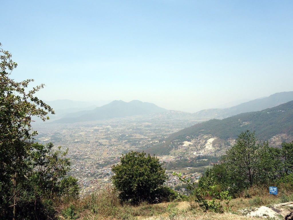 A panoramic view overlooking a valley with scattered buildings, surrounded by mountains under a hazy blue sky, with vegetation in the foreground.
