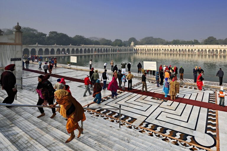 Visitors at a religious site, walking around an ornate pool with a long colonnade in the background, under a clear sky.