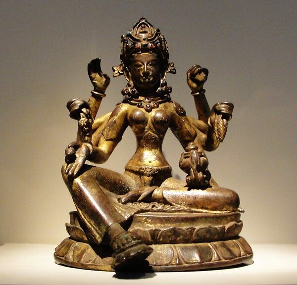 A bronze statue of a four-armed deity sitting in a meditative posture on a pedestal, displayed against a neutral background with spot lighting.