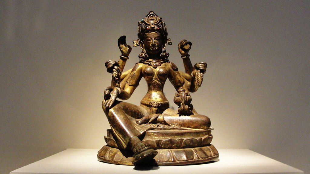 A bronze statue of a four-armed deity sitting in a meditative posture on a pedestal, displayed against a neutral background with spot lighting.