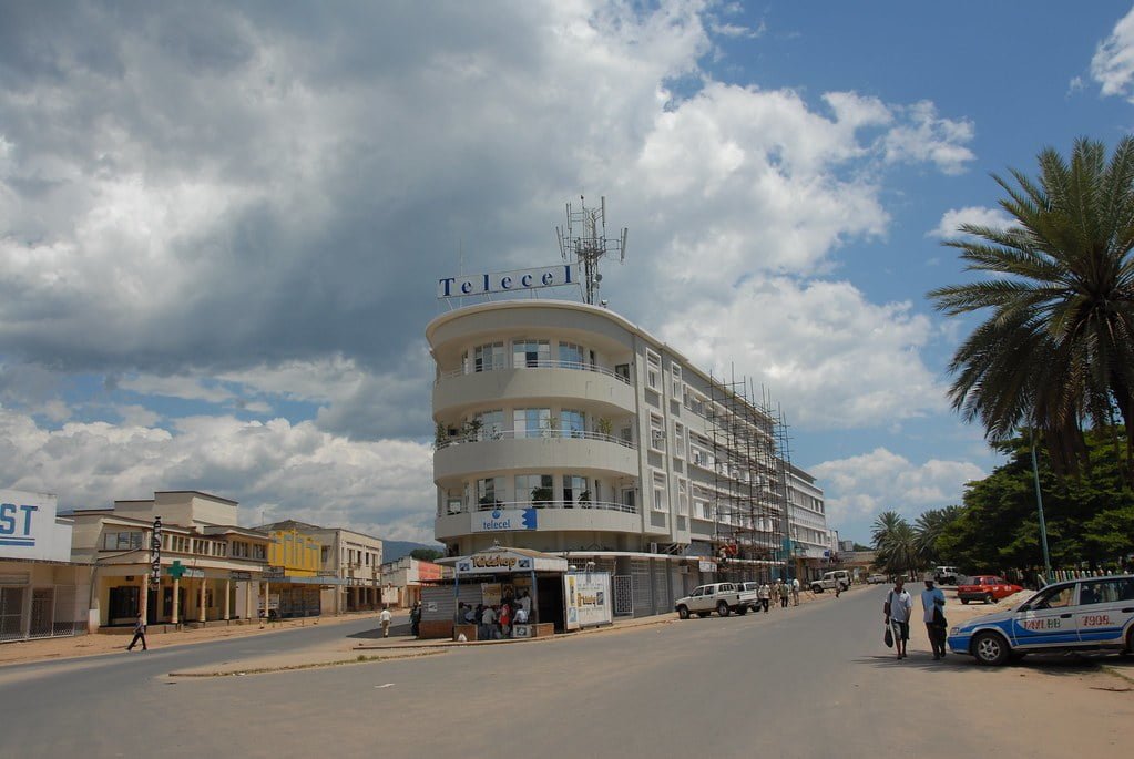 A street view in a town showing a multi-story corner building with "Telecel" signage, flanked by other buildings, palm trees, pedestrians, and parked cars under a cloudy sky.