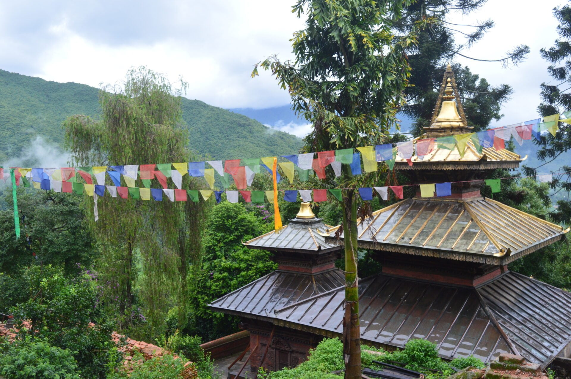 Traditional Nepalese pagoda-style temple roofs with golden spires, adorned with colorful prayer flags, set against a backdrop of lush green mountains shrouded in mist.