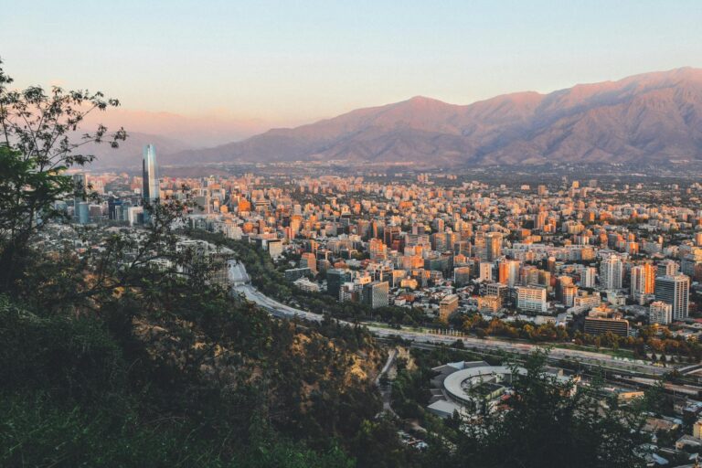 Aerial view of a city with dense buildings at sunset, framed by foliage in the foreground and mountains in the background, with a prominent skyscraper standing taller than its surroundings.