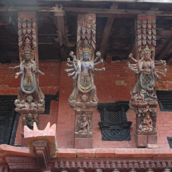 Three detailed wooden Hindu deity figures carved into the support beams of a traditional Nepalese building with ornate brickwork and dark wood window frames in the background.