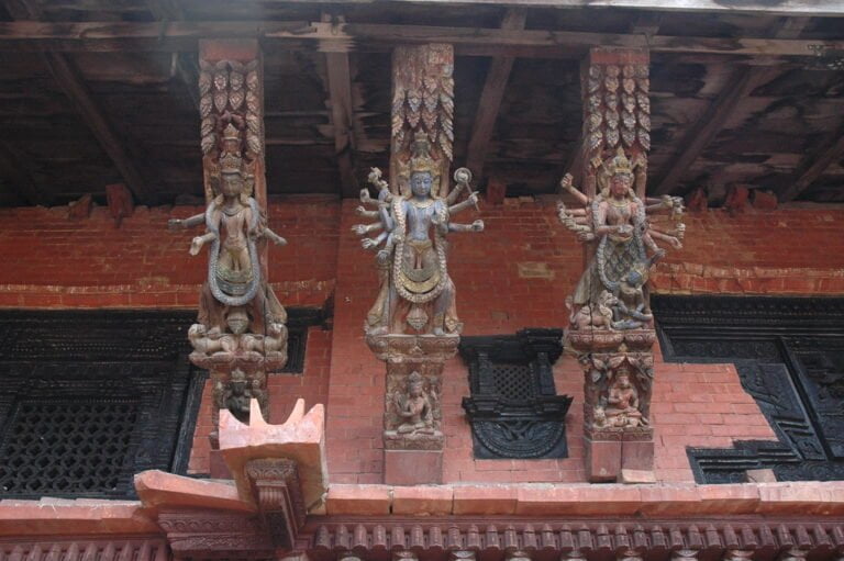 Three detailed wooden Hindu deity figures carved into the support beams of a traditional Nepalese building with ornate brickwork and dark wood window frames in the background.