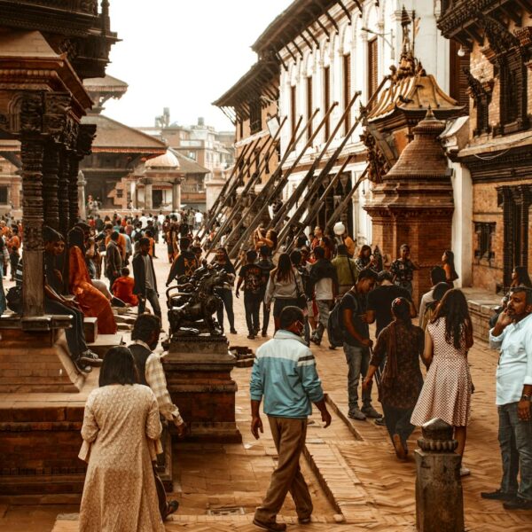 Alt text: A bustling scene at a traditional Nepalese square, with people walking, conversing, and sitting on the steps of ancient temples and structures under a warm, hazy sky.