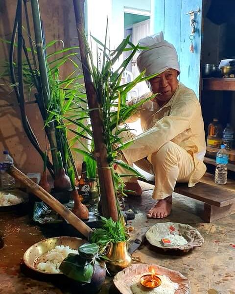 An elderly person in traditional attire sitting next to religious offerings, including plants, a lit lamp, and food items on leaf plates, inside a room with a rustic ambiance.