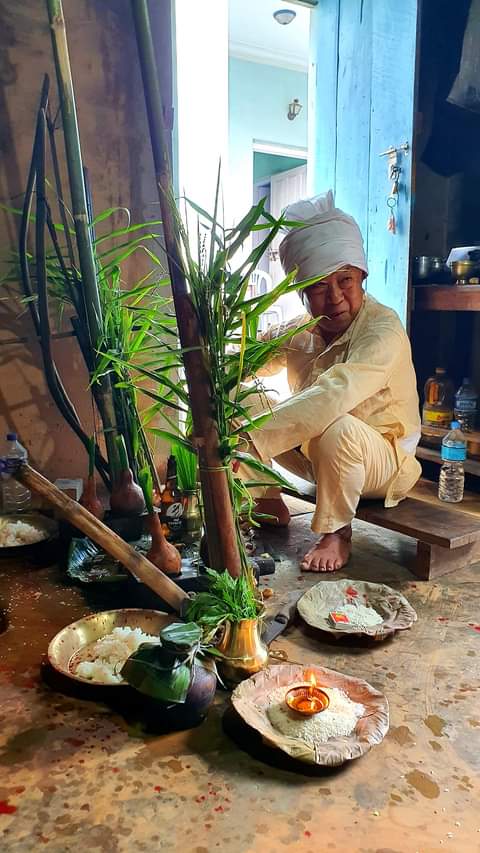 An elderly person in traditional attire sitting next to religious offerings, including plants, a lit lamp, and food items on leaf plates, inside a room with a rustic ambiance.