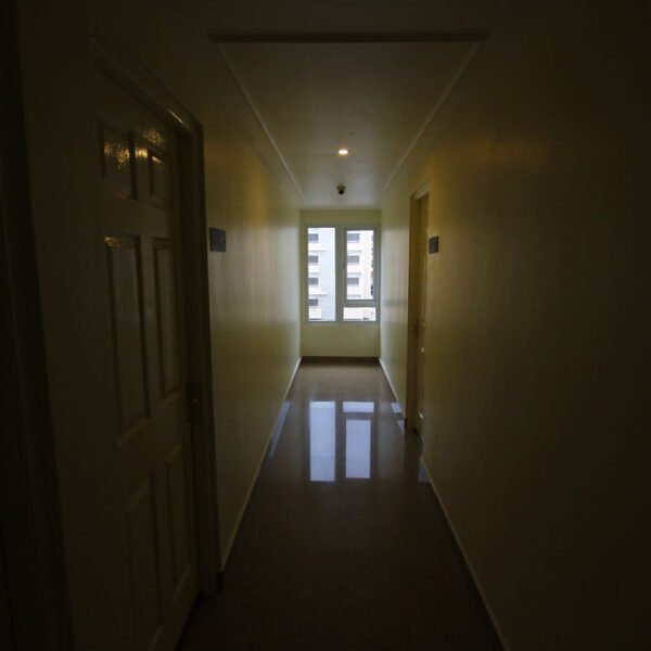A dimly lit hallway with closed doors on one side and a window at the far end letting in natural light.