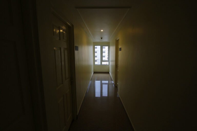 A dimly lit hallway with closed doors on one side and a window at the far end letting in natural light.