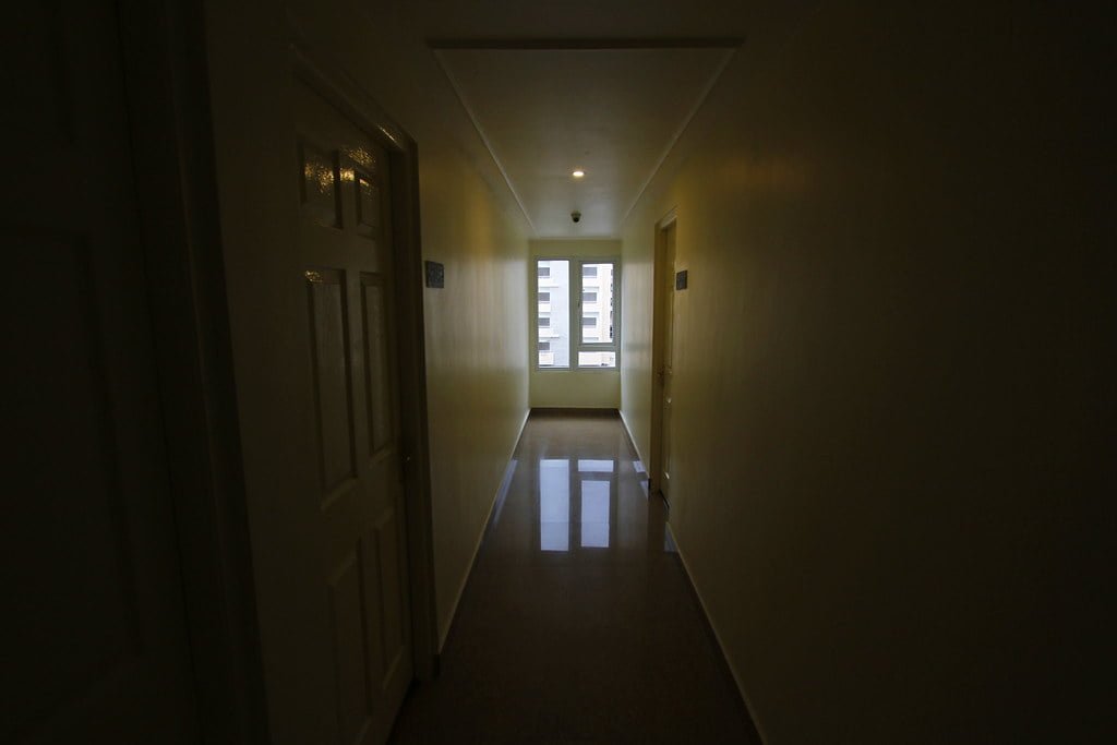 A dimly lit hallway with closed doors on one side and a window at the far end letting in natural light.