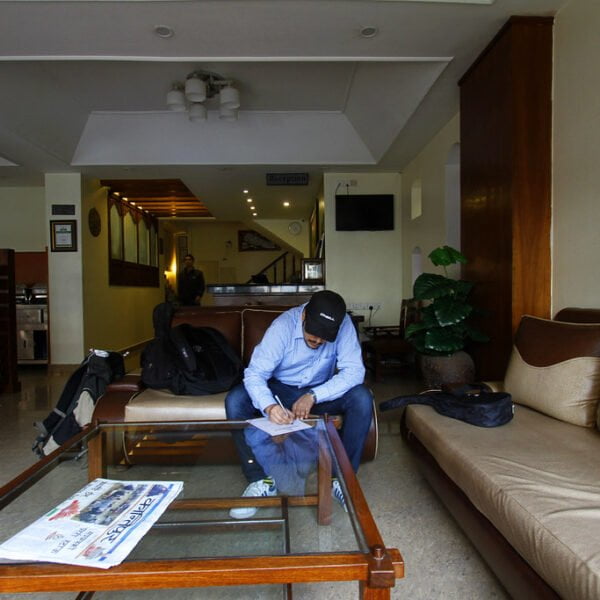A man wearing a cap and blue shirt is sitting in a hotel lobby, looking down at his phone with a bag next to him. The lobby has sofas, a wooden coffee table with a newspaper on it, and a reception counter in the background.