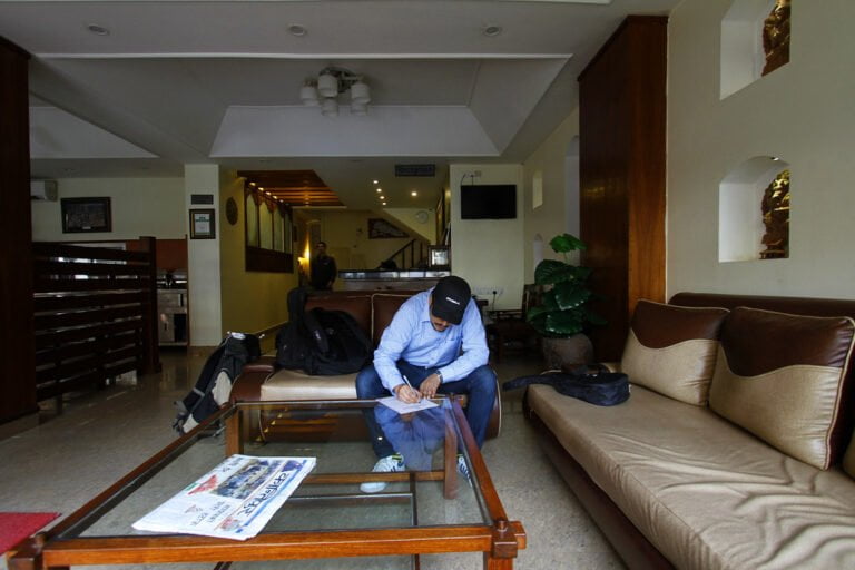 A man wearing a cap and blue shirt is sitting in a hotel lobby, looking down at his phone with a bag next to him. The lobby has sofas, a wooden coffee table with a newspaper on it, and a reception counter in the background.