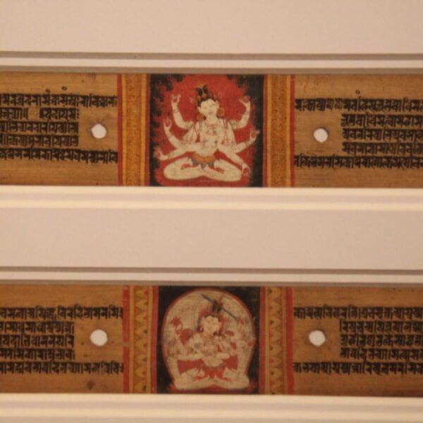 Two framed sections of an ancient manuscript with Sanskrit text and colorful illustrations of Hindu deities in the center, displayed on a white wall.