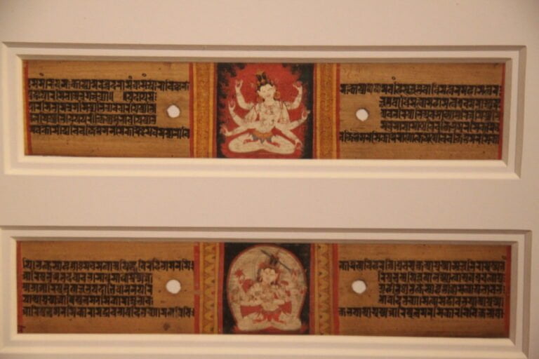 Two framed sections of an ancient manuscript with Sanskrit text and colorful illustrations of Hindu deities in the center, displayed on a white wall.