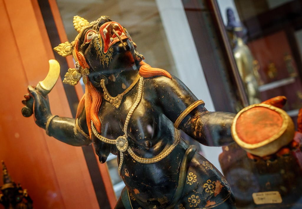 A detailed statue of a Hindu deity with dark blue skin, multiple arms, and ornate gold and red accents displayed behind a glass enclosure.