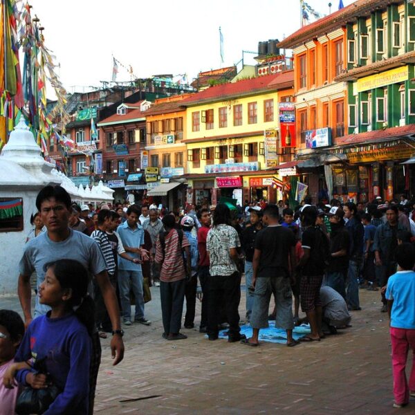 A bustling street scene with people gathered in a market area, featuring colorful traditional buildings and prayer flags, capturing the vibrant atmosphere of a Nepalese town square.