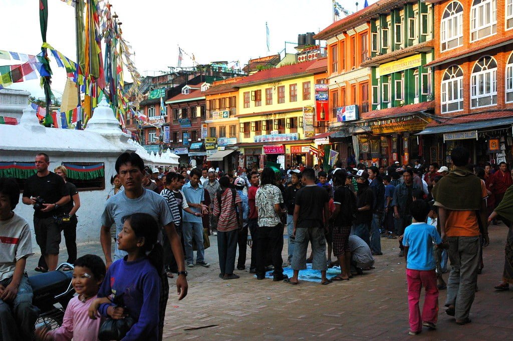 A crowded market square with people walking around and talking, colorful buildings with shopfronts in the background, prayer flags, and a stupa in the foreground.