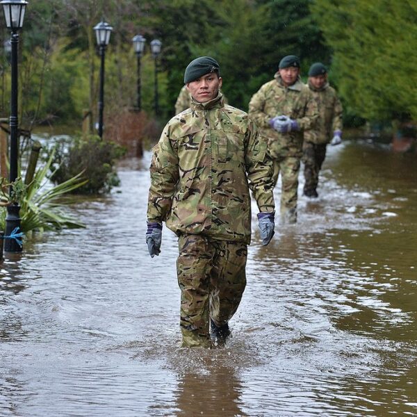 Soldiers in camouflage uniforms wading through a flooded street with water submerging the base of street lamps and vegetation.