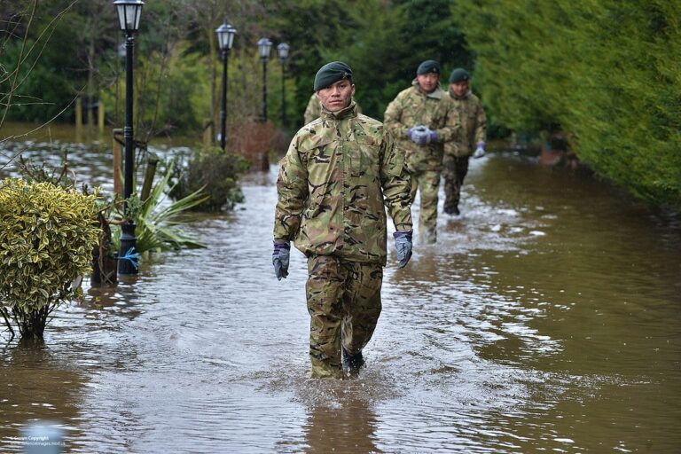 Soldiers in camouflage uniforms wading through a flooded street with water submerging the base of street lamps and vegetation.