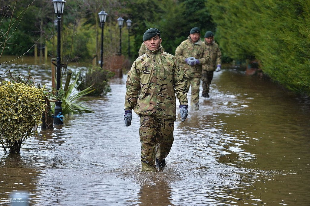 Soldiers in camouflage uniforms wading through a flooded street with water submerging the base of street lamps and vegetation.