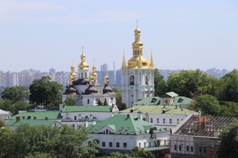 A view of a historic monastery featuring black and white structures with golden domes, set against a modern city skyline in the background and construction work in the foreground.