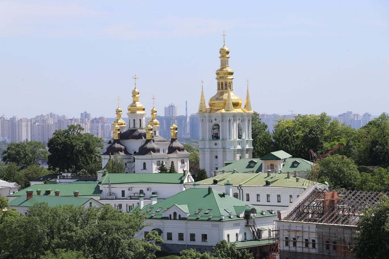 A view of a historic monastery featuring black and white structures with golden domes, set against a modern city skyline in the background and construction work in the foreground.