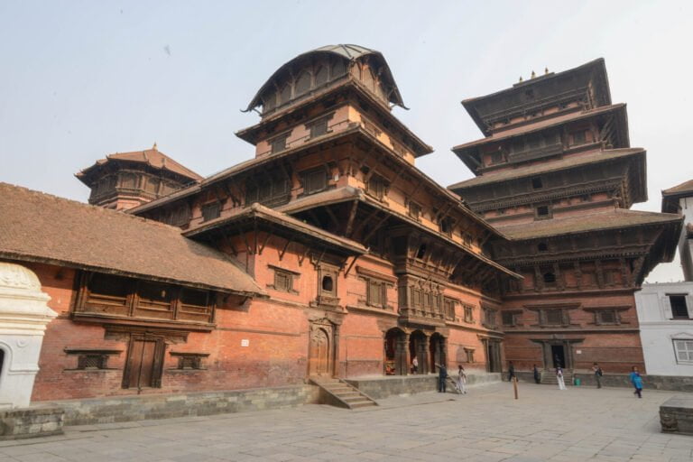 Alt text: A traditional Nepalese temple complex with multi-tiered pagoda-style architecture, featuring ornate wooden carvings and brickwork, with a few people walking in the courtyard.