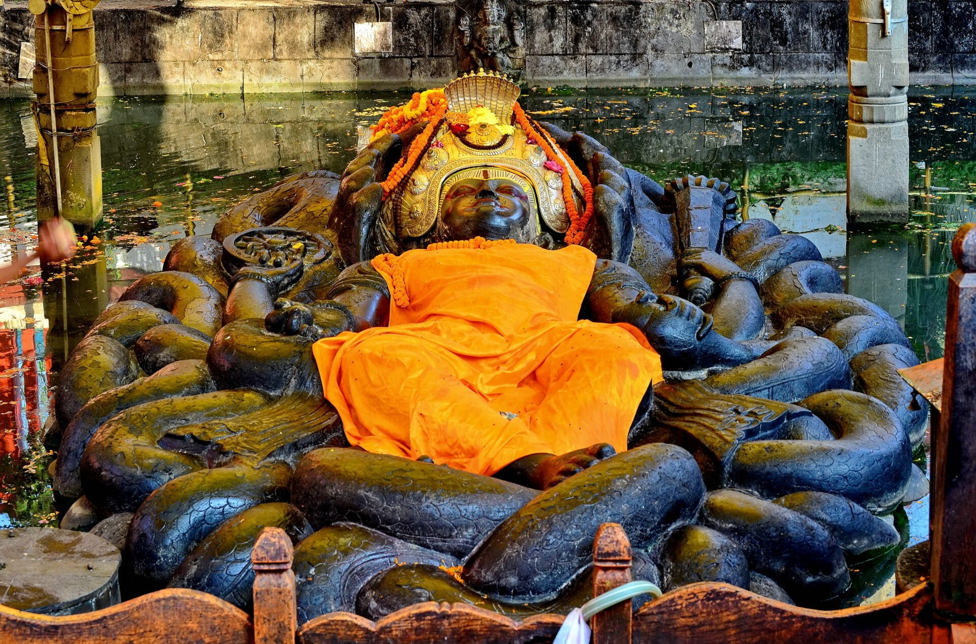 A statue of a Hindu deity reclining on a serpent, adorned with orange cloth and floral garlands, partially submerged in water with leaves floating on the surface.