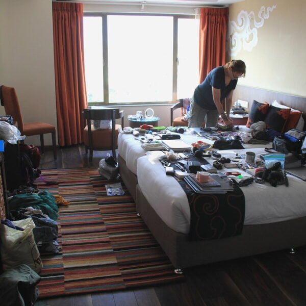 A person standing and organizing various items spread out on a bed in a messy hotel room with an open suitcase and clutter on the floor and furniture.