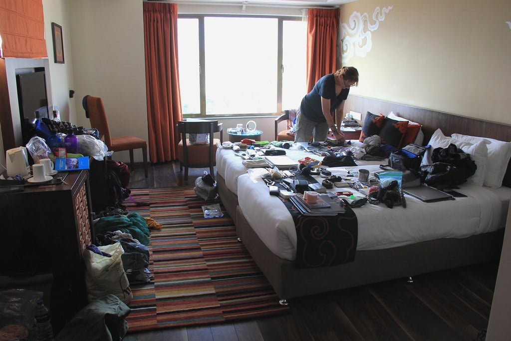 A person standing and organizing various items spread out on a bed in a messy hotel room with an open suitcase and clutter on the floor and furniture.