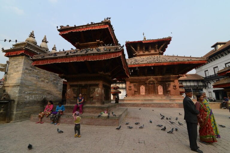 Two multi-tiered pagoda-style temples with ornate wood carvings dominate the center of a dusty brick square. Local people are sitting and standing around; pigeons scatter on the ground. A woman in traditional clothing converses with a man in a suit and hat.