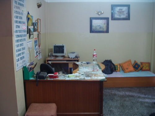 A small, cluttered room with a computer desk and various items on it, a bed with colorful pillows to the right, a carpet on the floor, and framed pictures on the wall.