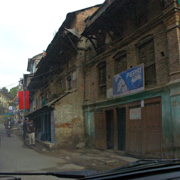 View from a car showing a worn, brick building with a Pepsi advertisement on its facade, a street with people, bikes, and parked vehicles, and colorful flags strung above in a developing urban area.