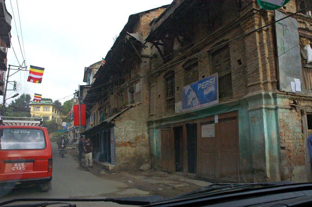 View from a car showing a worn, brick building with a Pepsi advertisement on its facade, a street with people, bikes, and parked vehicles, and colorful flags strung above in a developing urban area.