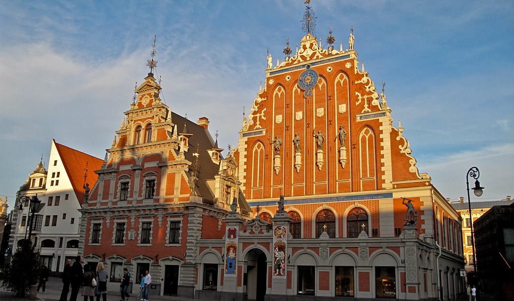 The House of the Blackheads and another historical building bathed in warm sunlight against a blue sky, located in a European city square with a few pedestrians nearby.